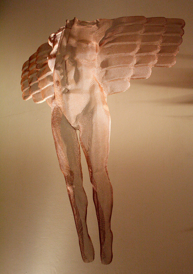 A transparent bronze angel suspended between trees at a sculpture garden exhibition